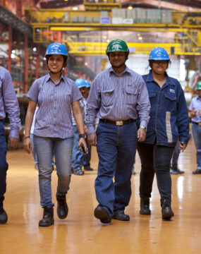 A group of five workers in a steel plant are walking in line heading towards the camera. They are wearing jeans and hards hats.