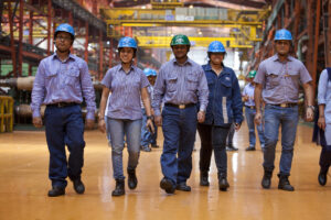 A group of five workers in a steel plant are walking in line heading towards the camera. They are wearing jeans and hards hats.