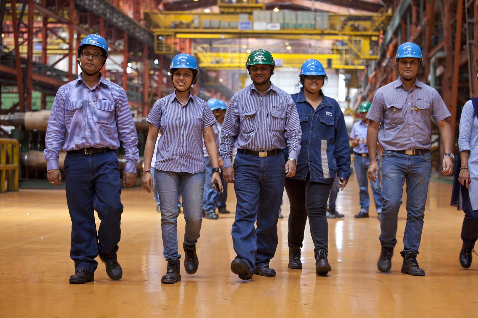 A group of five workers in a steel plant are walking in line heading towards the camera. They are wearing jeans and hards hats.