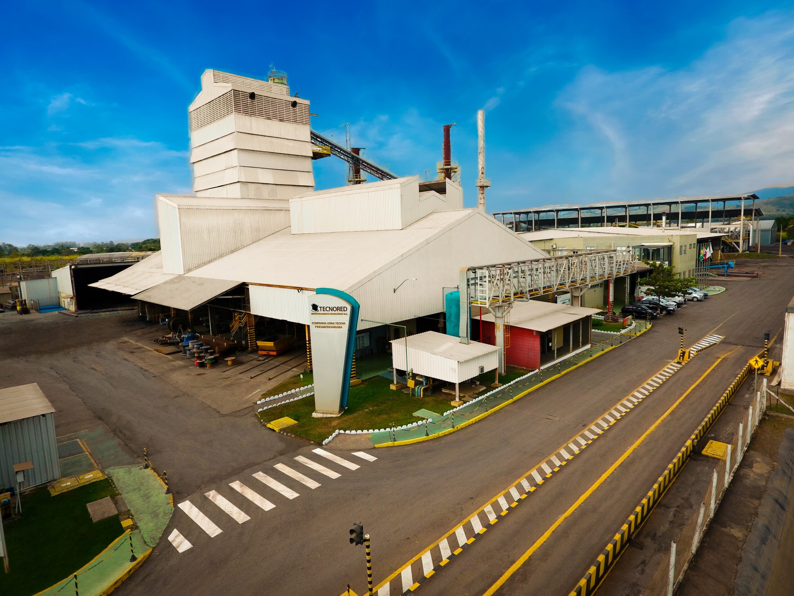 An industrial plant in Brazil. The buildings are white against a blue sky. A road runs alongside the factory.